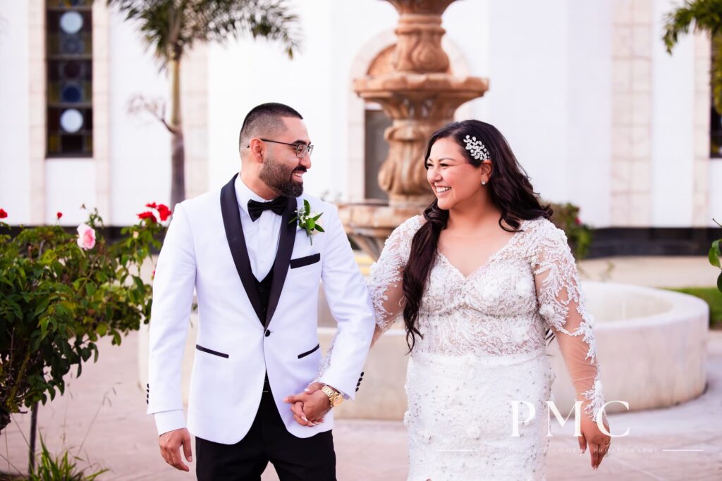 A bride and groom smile and walk hand-in-hand in front of a church fountain on their San Diego wedding day.