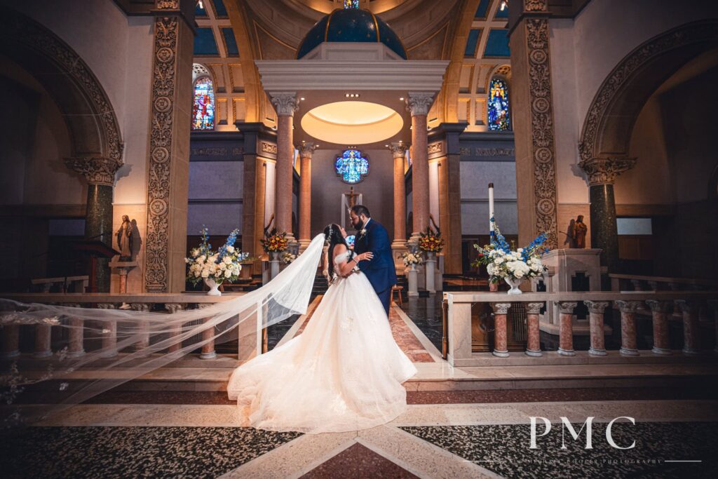 A portrait of a groom and a bride with a sweeping cathedral veil embrace each other on the altar of the Immaculata Church at the University of San Diego.