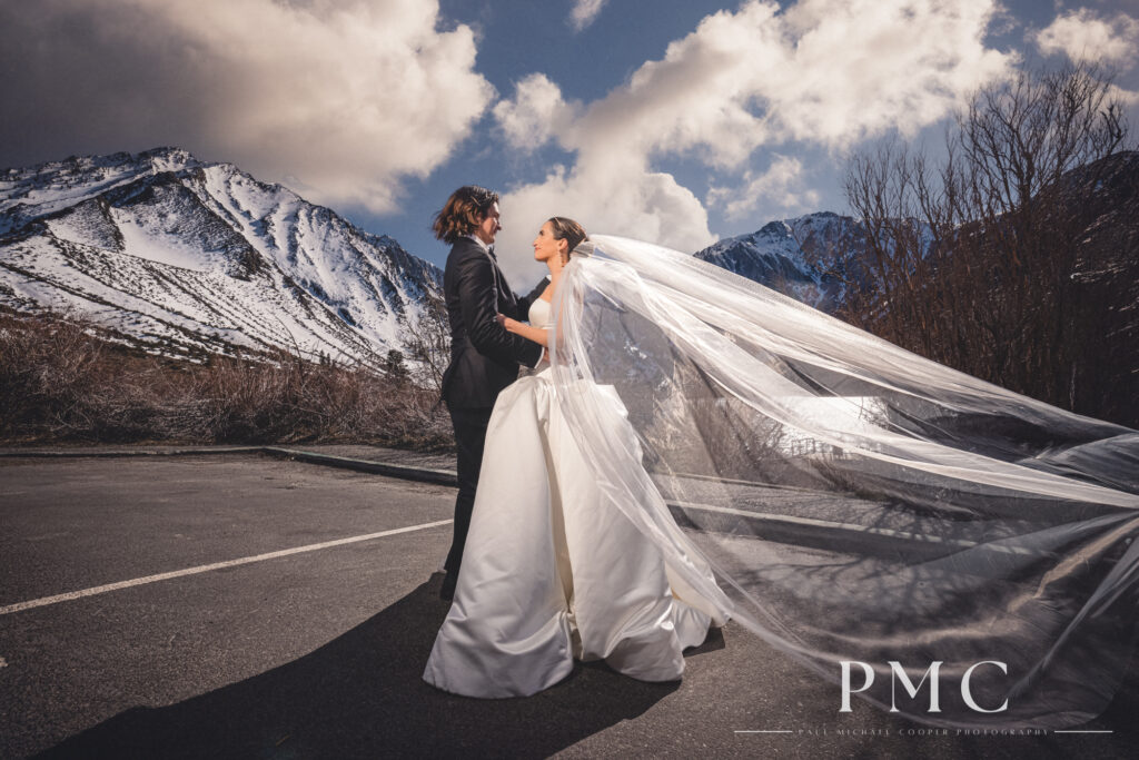 A bride with a sweeping veil embraces her groom on their wedding day in front of a mountain landscape.