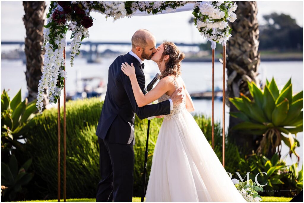 A bride and groom kiss at their ceremony at a waterfront wedding venue with a view of Mission Bay.