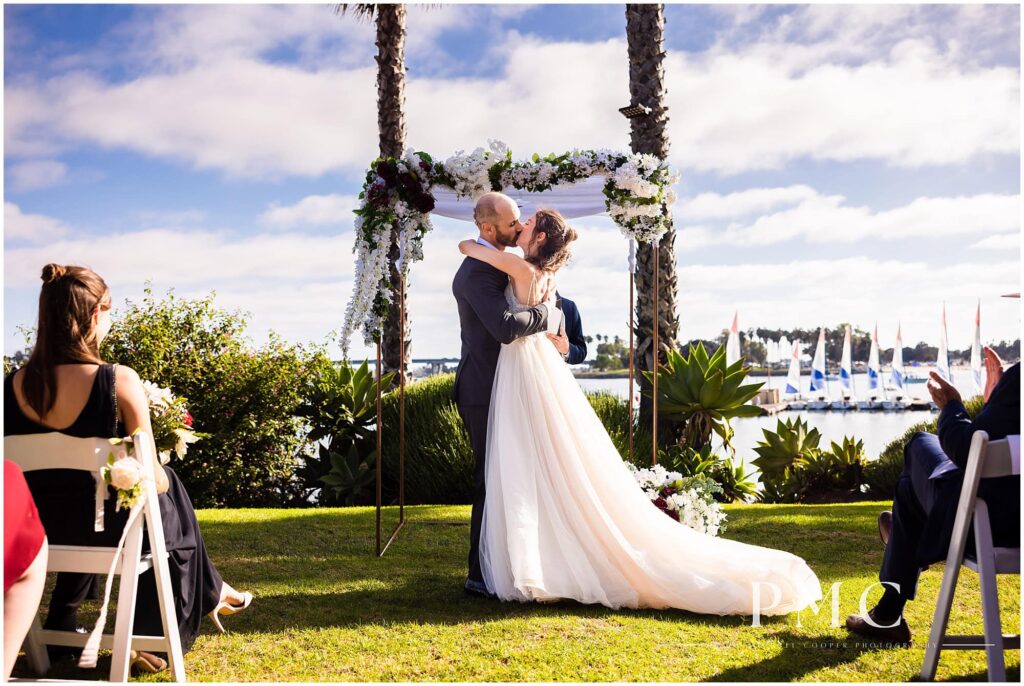 A bride and groom kiss at their ceremony at a waterfront wedding venue with a view of Mission Bay.