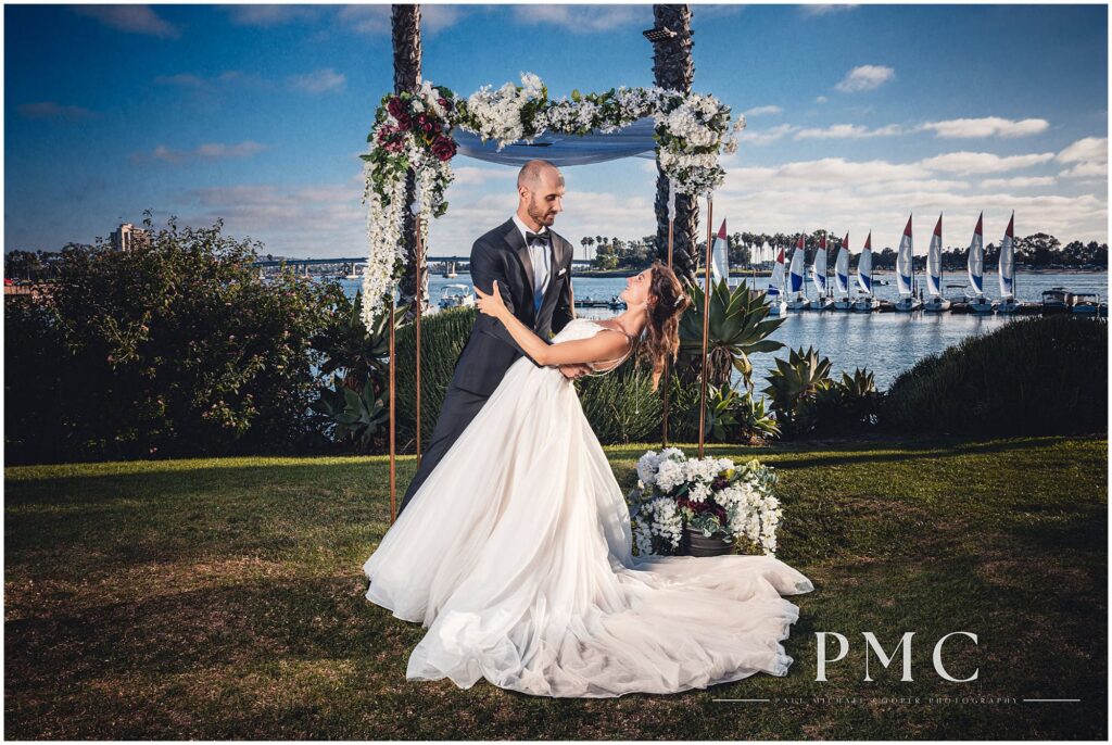 A bride and groom share a dramatic 
bayside dip in front of their floral arch at their waterfront wedding venue with a view of Mission Bay.