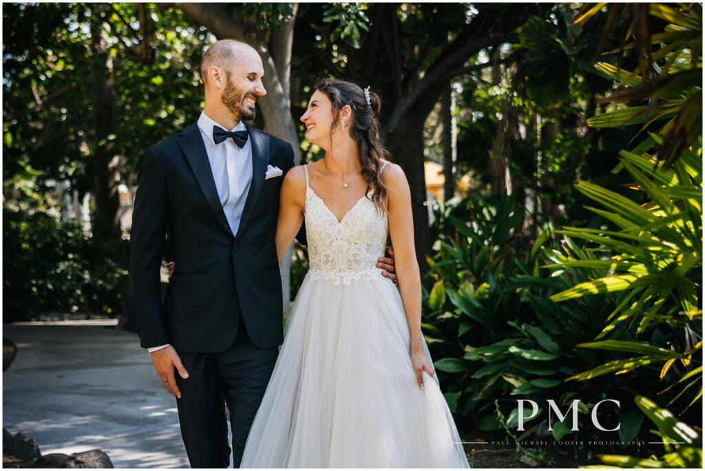 A bride and groom smile as they walk together through the gardens at Paradise Point Resort & Spa in Mission Bay.