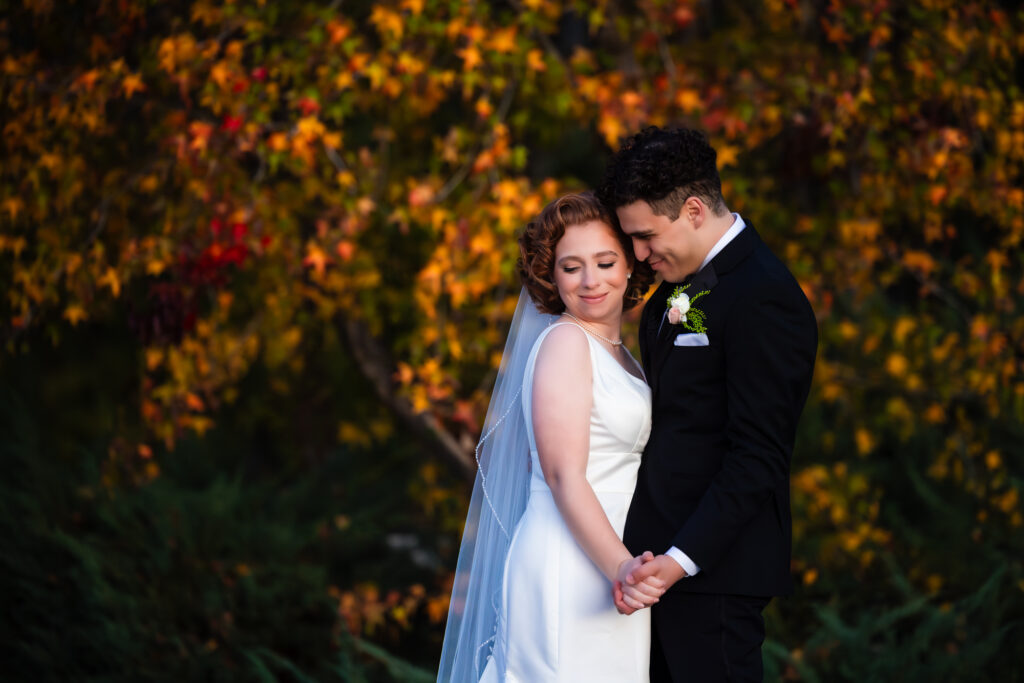 A bride and groom holding hands and smiling in front of a tree with colorful fall and autumn leaves.