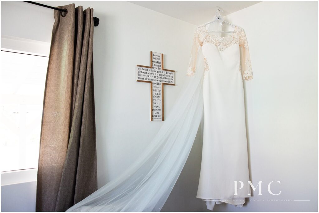 A bride's wedding dress and veil hang from a ceiling hook beside a custom cross.