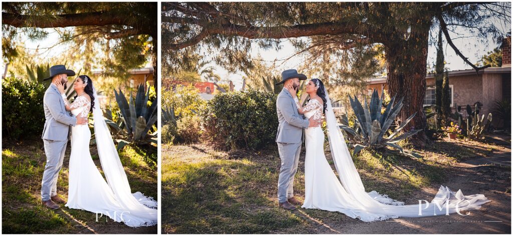 A bride with a long, flowing train and veil and elegant hairpiece embraces her groom, who is wearing a grey suit with cowboy boots and a cowboy hat on their country-style wedding day.