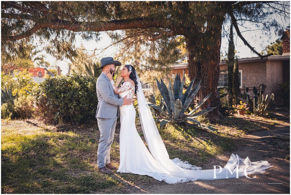 A bride with a long, flowing train and veil and elegant hairpiece embraces her groom, who is wearing a grey suit with cowboy boots and a cowboy hat on their country-style wedding day.