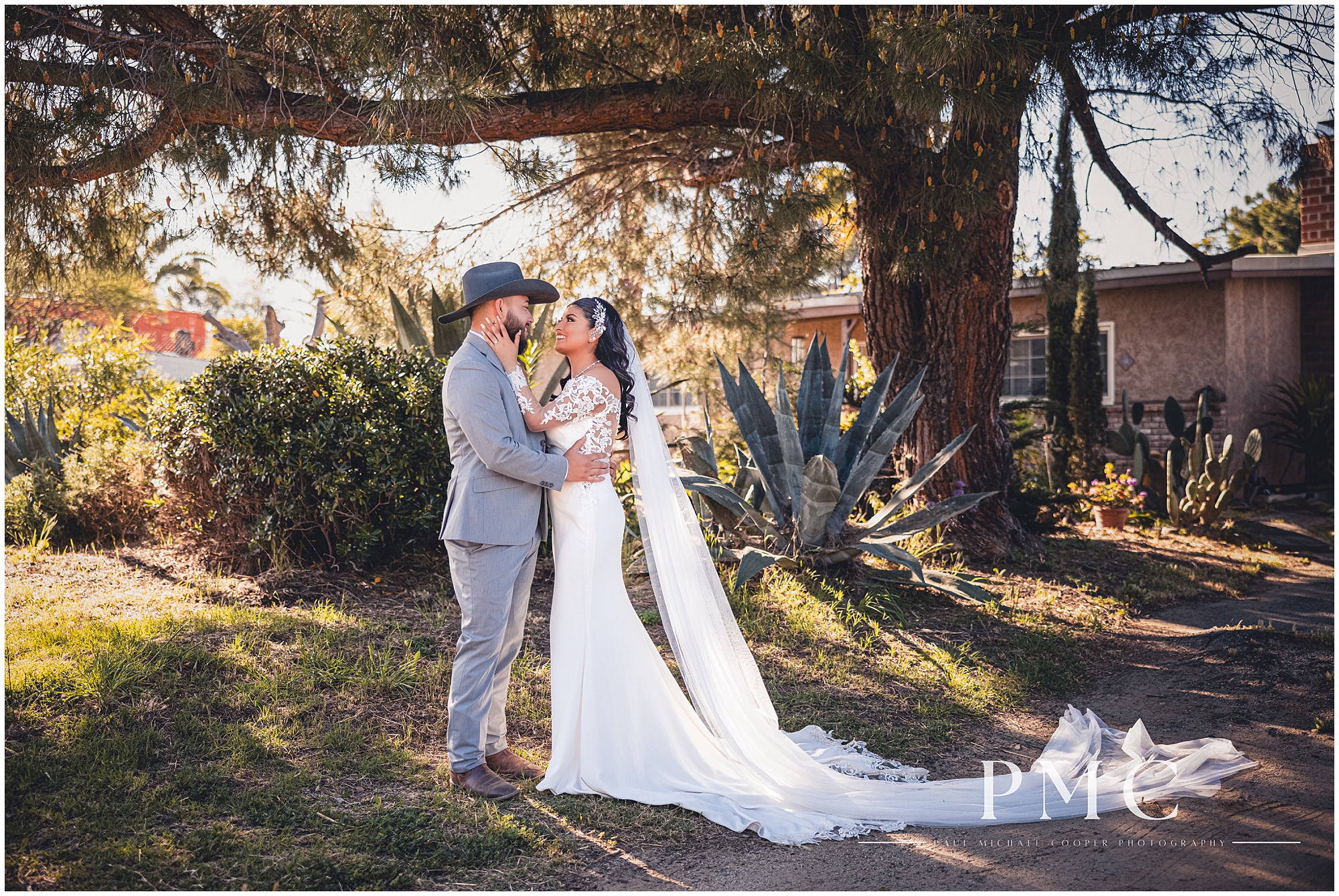 A bride with a flowing train and veil embraces her groom in a suit with boots and a cowboy hat in a nature scene.