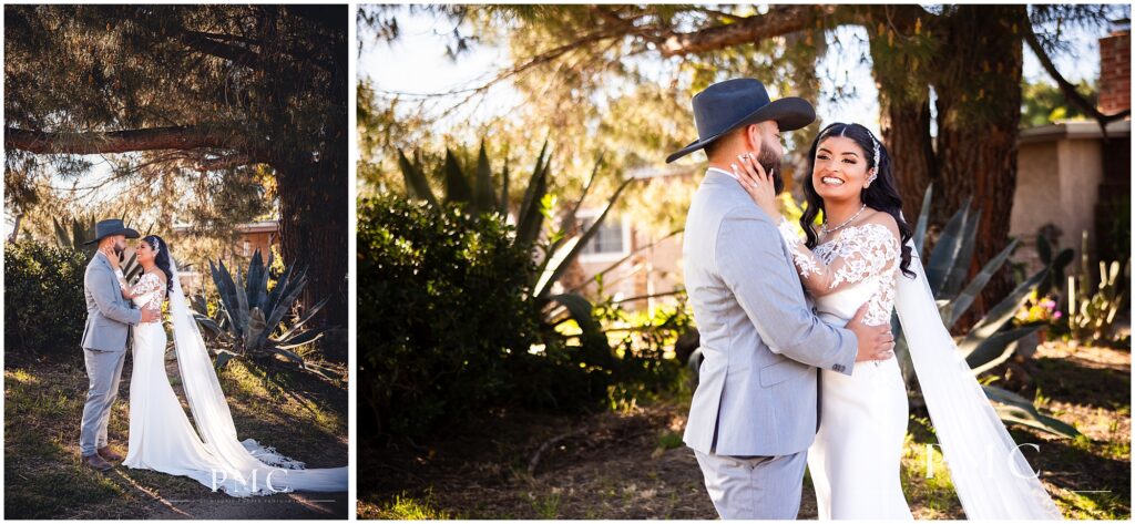 A bride with a long, flowing train and veil and elegant hairpiece embraces her groom, who is wearing a grey suit with cowboy boots and a cowboy hat on their country-style wedding day.