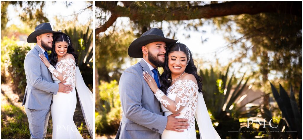 A bride with a long, flowing train and veil and elegant hairpiece embraces her groom, who is wearing a grey suit with cowboy boots and a cowboy hat on their country-style wedding day.