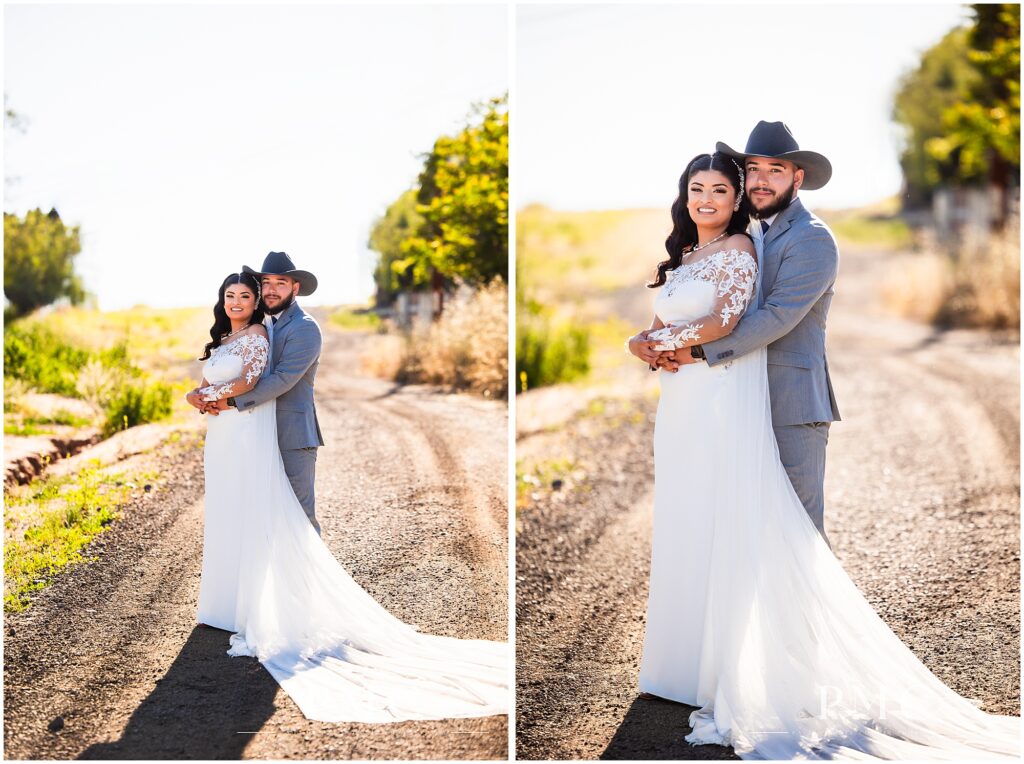 A bride with a long, flowing train and veil and elegant hairpiece embraces her groom, who is wearing a grey suit with cowboy boots and a cowboy hat on their country-style wedding day.