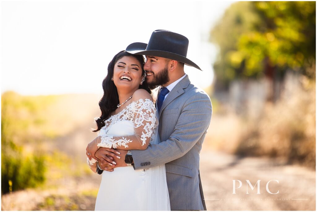 A groom wearing a cowboy hat embraces his bride and laughs with her.