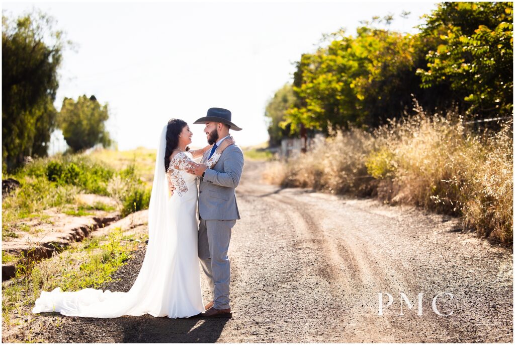 A bride with a long, flowing train and veil and elegant hairpiece dances on a rustic dirt road with her groom, who is wearing a grey suit with cowboy boots and a cowboy hat on their country-style wedding day.