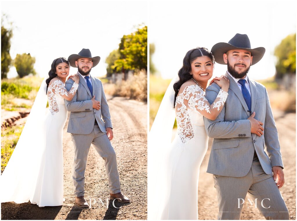 A bride with a long, flowing train and veil and elegant hairpiece poses on a rustic dirt road with her groom, who is wearing a grey suit with cowboy boots and a cowboy hat on their country-style wedding day.