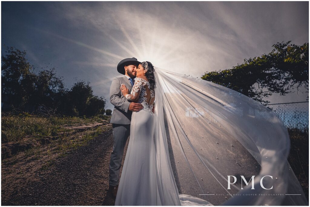 A bride with a long, flowing train and veil and elegant hairpiece dances on a rustic dirt road with her groom, who is wearing a grey suit with cowboy boots and a cowboy hat on their country-style wedding day.