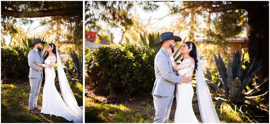 A bride with a long, flowing train and veil and elegant hairpiece embraces her groom, who is wearing a grey suit with cowboy boots and a cowboy hat on their country-style wedding day.