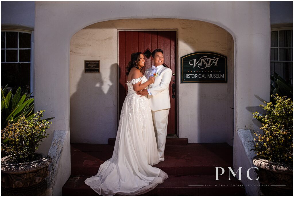 Two brides embrace and smile on the front steps of the terracotta Rancho Minerva Historical Museum Wedding Venue in Vista.