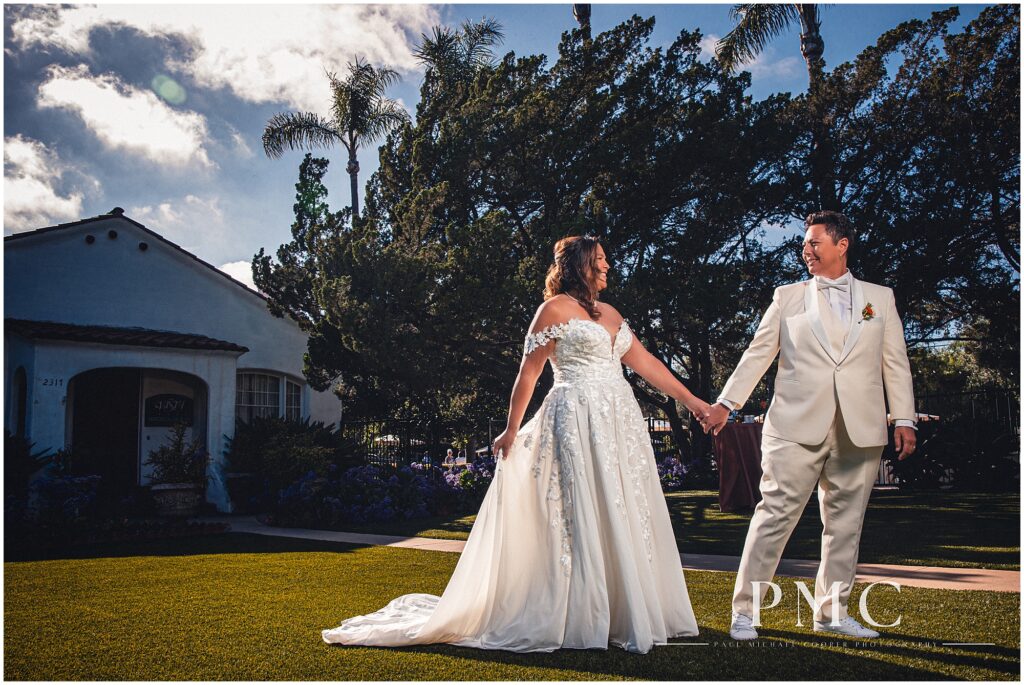 Two brides walk hand-in-hand on the front lawn in front of the main terracotta building at the historical Rancho Minerva Venue.