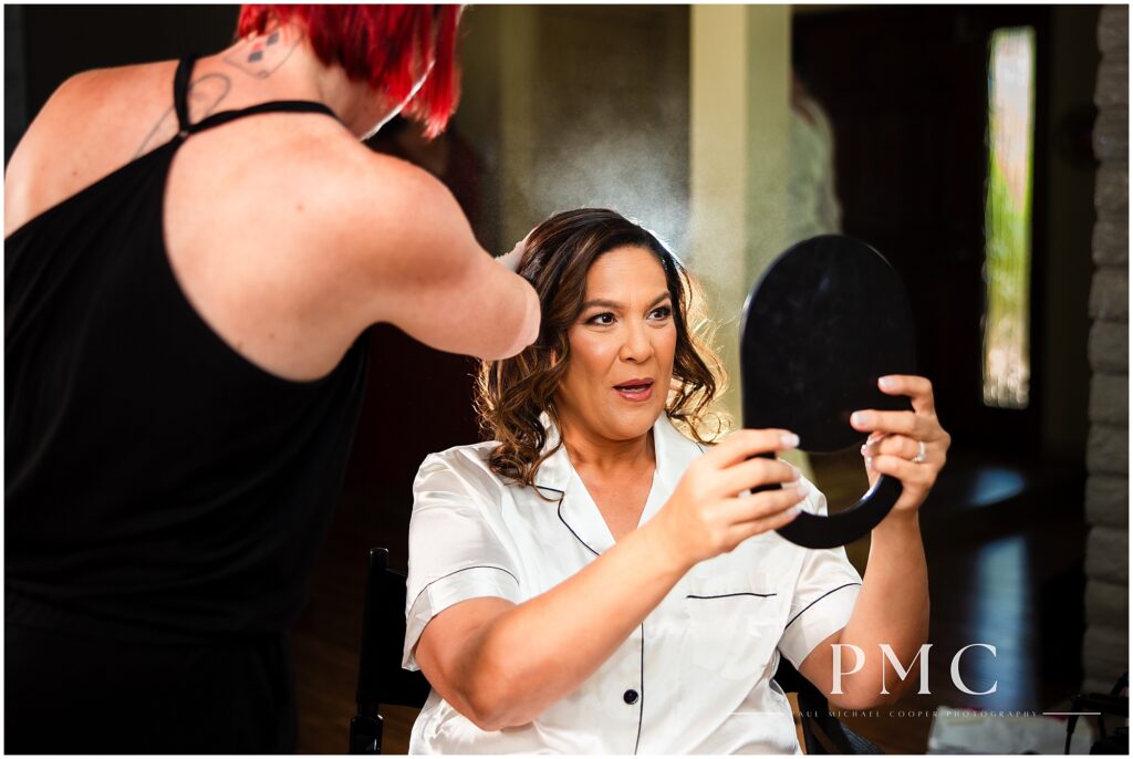 A bride gets her hair done while she looks in the mirror on her summer wedding day in Vista.