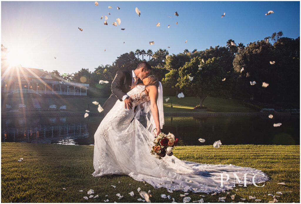 A bride and groom share a romantic, sunlit dip and kiss as flower petals float around them on the edge of the heart-shaped lake at Grand Tradition Estate & Gardens in Fallbrook, Southern California.