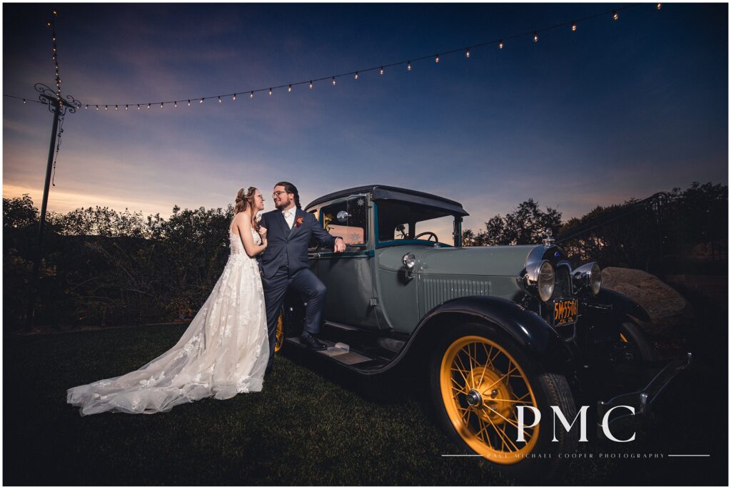 A bride and groom share a smile as they pose next to a vintage classic car below market lights at sunset on their fall wedding day in Escondido.