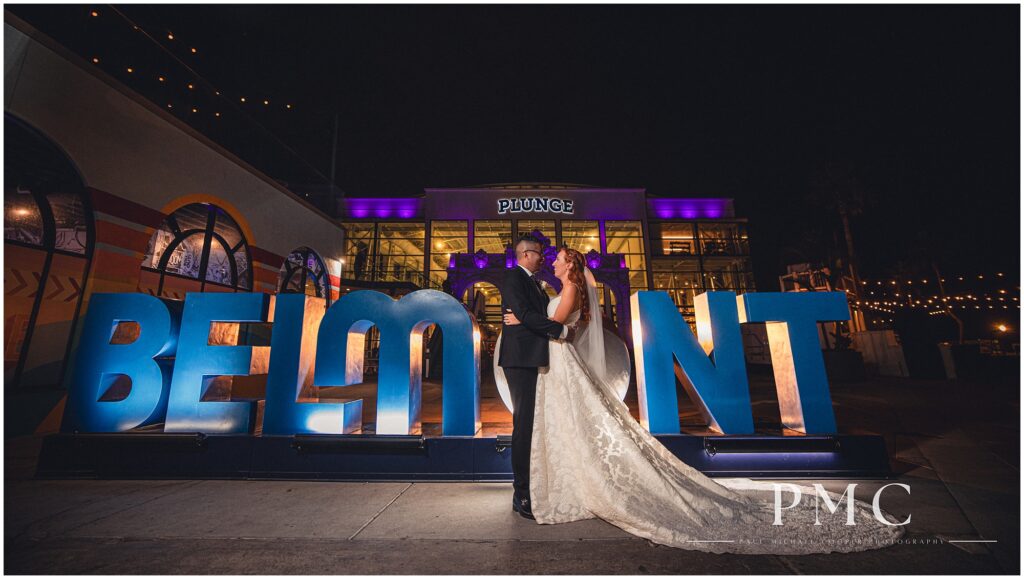 A bride and groom share a dramatic embrace in a cinematic night portrait in front of the main sign at Belmont Park, where they held their wedding, summer 2024.