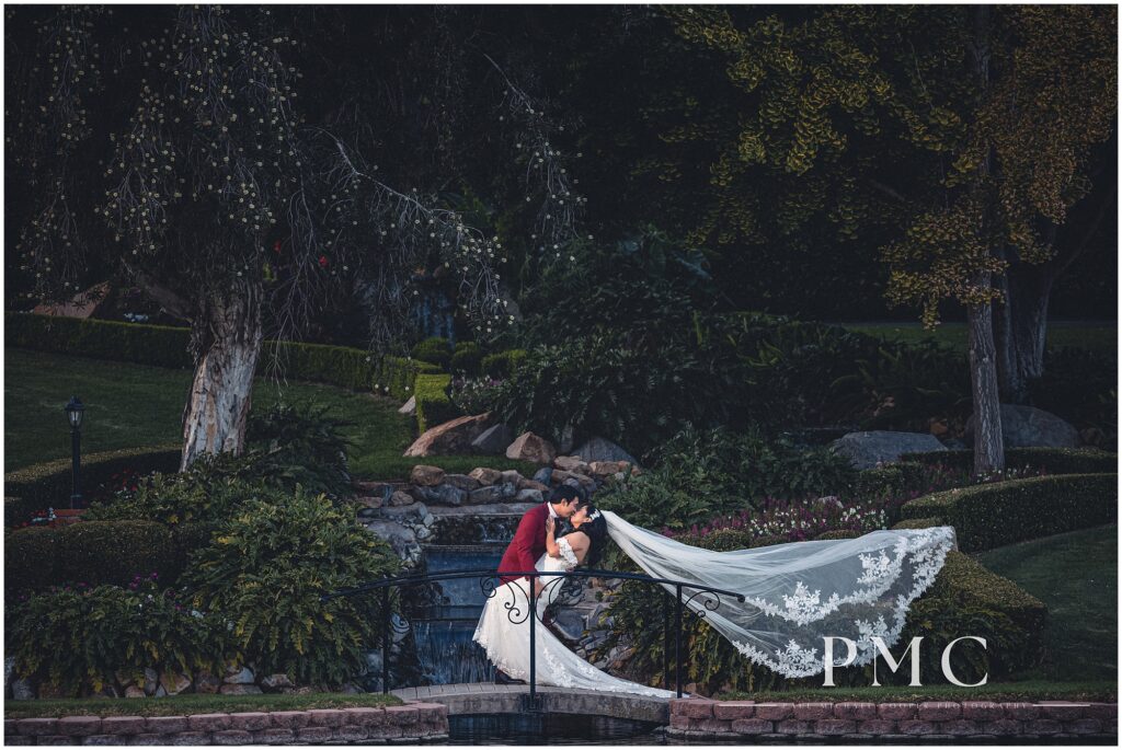 A bride with a sweeping veil and a groom in a colorful, burgundy suit share a romantic dip kiss on a bridge in front of a waterfall on their fall wedding day at Grand Tradition Estate & Gardens in Fallbrook, Southern California.