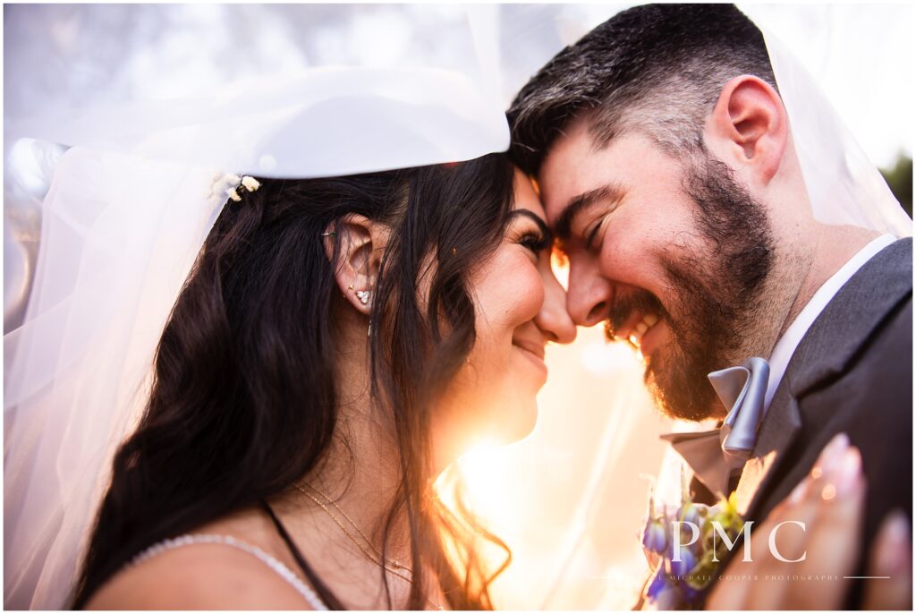 A bride and groom smile and touch noses under the bride's veil in a golden, sunlit moment on their wedding day at Los Willows Wedding Venue in Fallbrook.