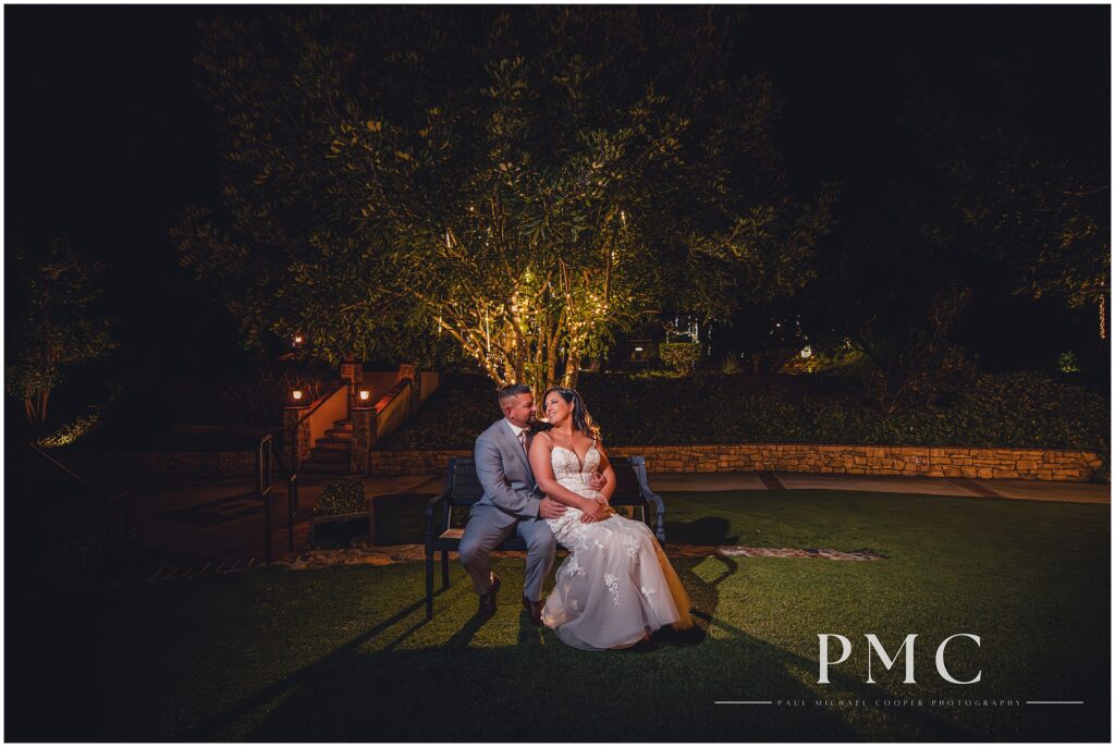 A bride and groom smile at each other while seated on a bench in a dramatic night portrait on their wedding day at Los Willows Wedding Venue, February 2024.