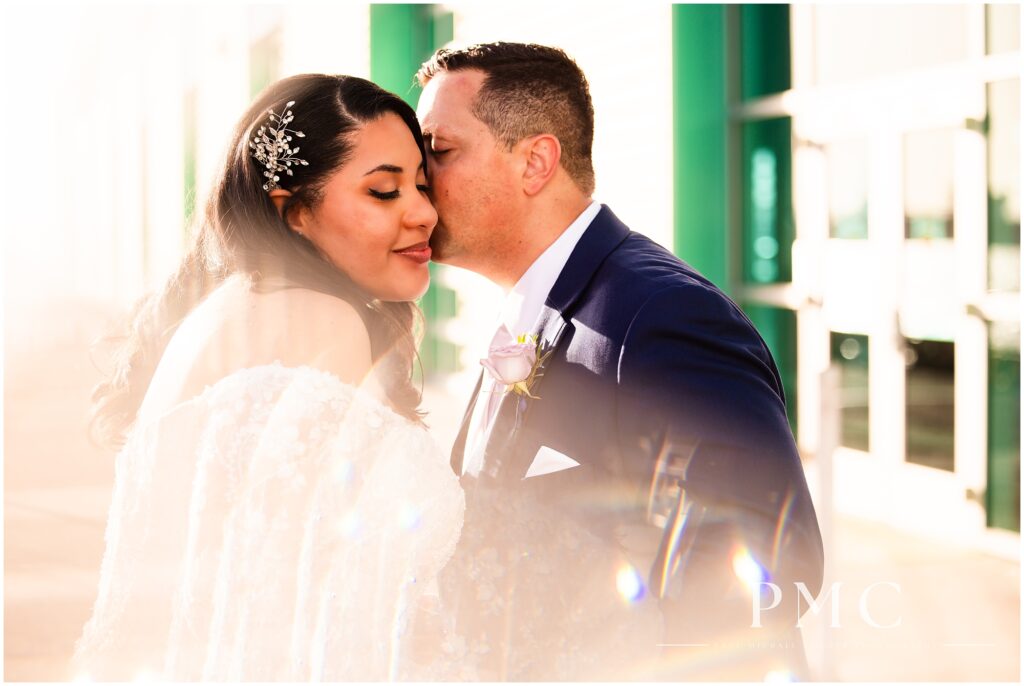 A groom kisses his bride on the cheek in a romantic, golden hour moment on the Broadway Pier in San Diego.