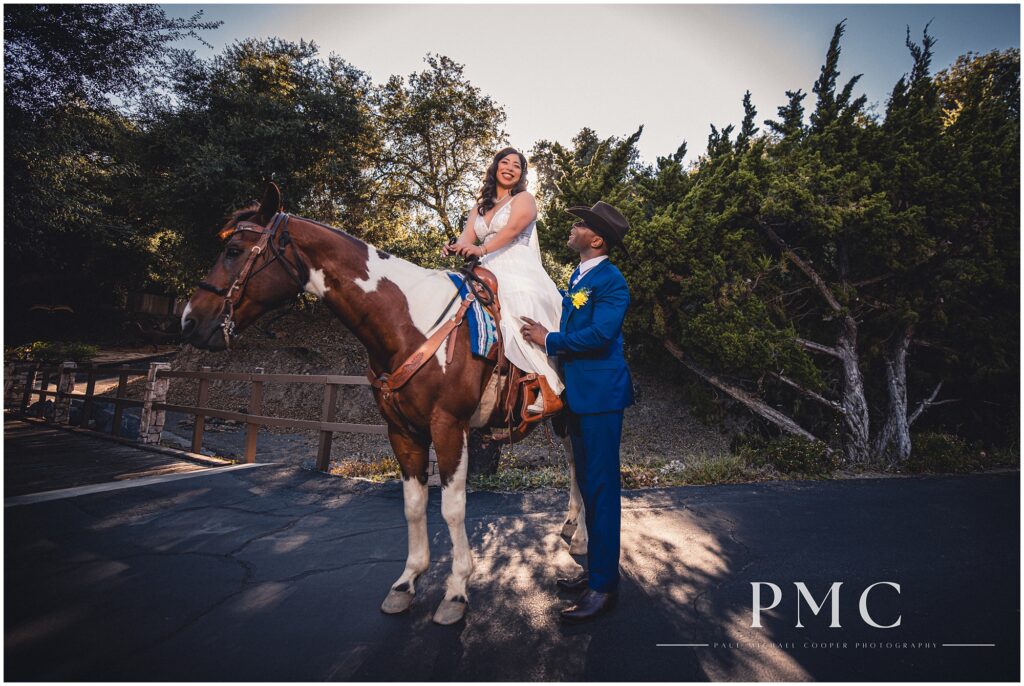 A bride sits atop a horse, while her groom stands beside them wearing a cowboy hat and blue suit on their wedding day at Los Willows Wedding Venue in Fallbrook, Southern California.