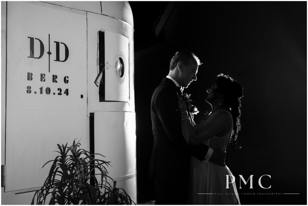 A dramatic black-and-white photo of a couple embracing by their custom monogram on the day of their 25th anniversary wedding vow renewal.