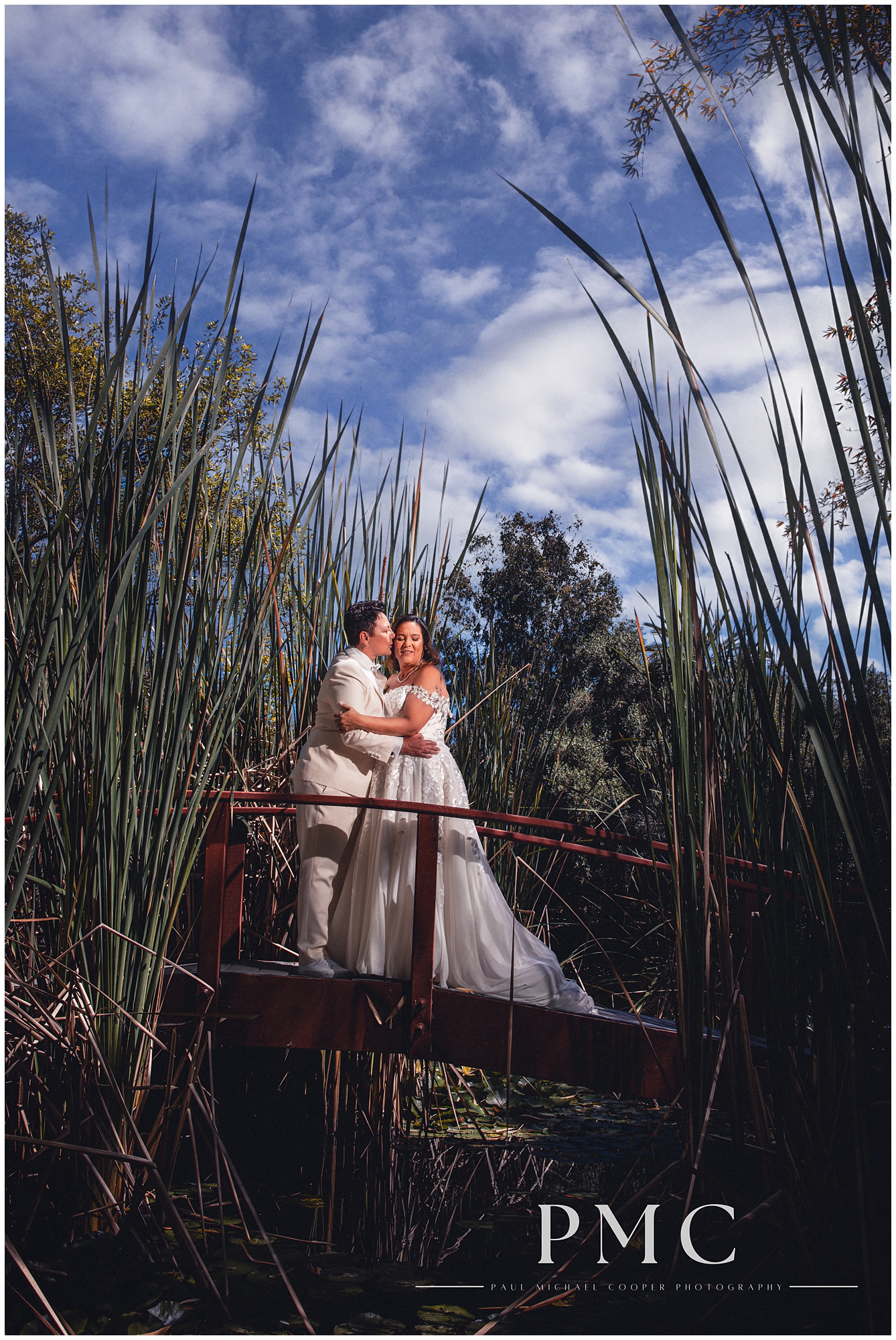 Two brides share a romantic kiss on a bridge in nature on their summer wedding day in Vista, Southern California.