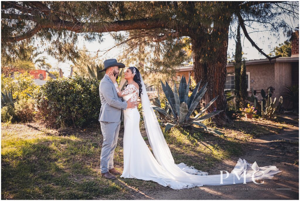 A bride with a long train and veil and groom wearing a cowboy hat smile and embrace each other surrounded by nature on their wedding day in Ramona, Southern California.