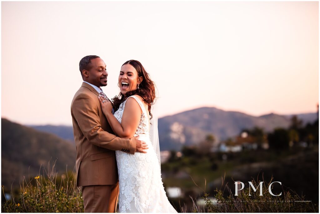 A bride has a big laugh as her groom embraces her in their nature sunset portraits at Tivoli Wedding Venue in Fallbrook, Southern California.