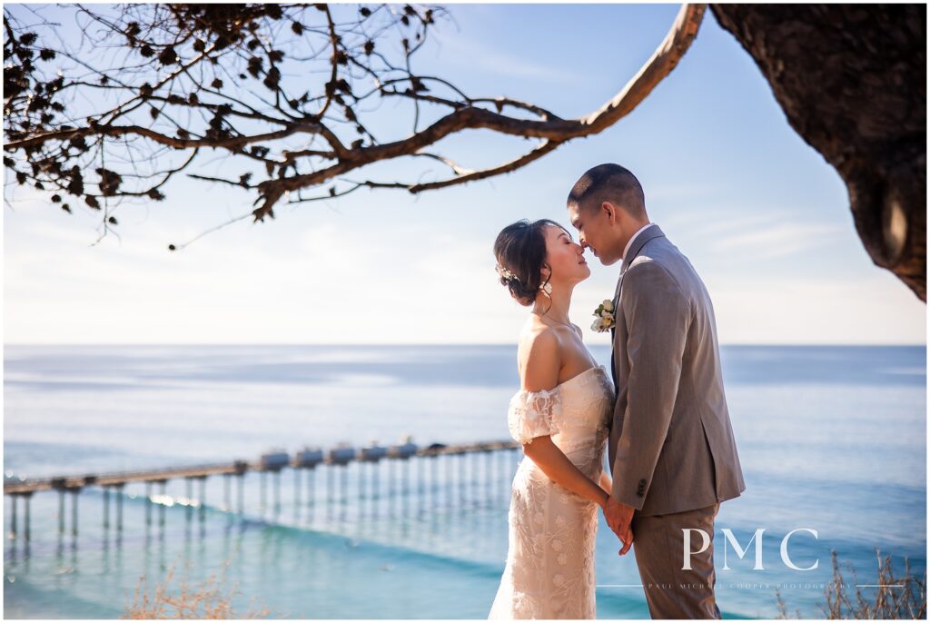 A bride and groom touch noses and hold hands in an intimate moment overlooking the Scripps Pier on their wedding day at the Martin Johnson House in La Jolla, San Diego.