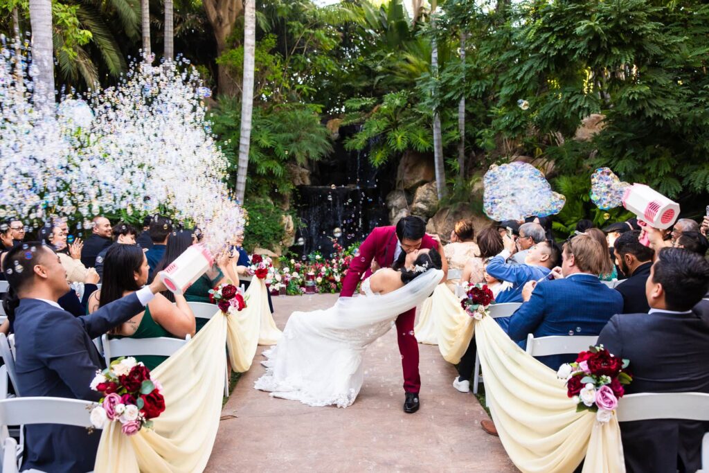 A bride and groom share a dip kiss as they recess down the aisle at the end of their wedding ceremony Grand Tradition Estate in Fallbrook. Friends with bubble guns shower them with bubbles as they recess. Photo by PMC Photography.