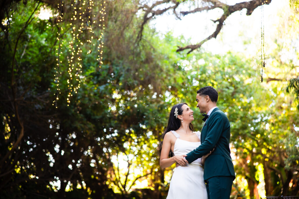 A bride and a groom in a green suit embrace each other under tall oak trees at Green Gables Wedding Estate, a Southern California wedding venue in San Marcos, owned by Trademark Venues.