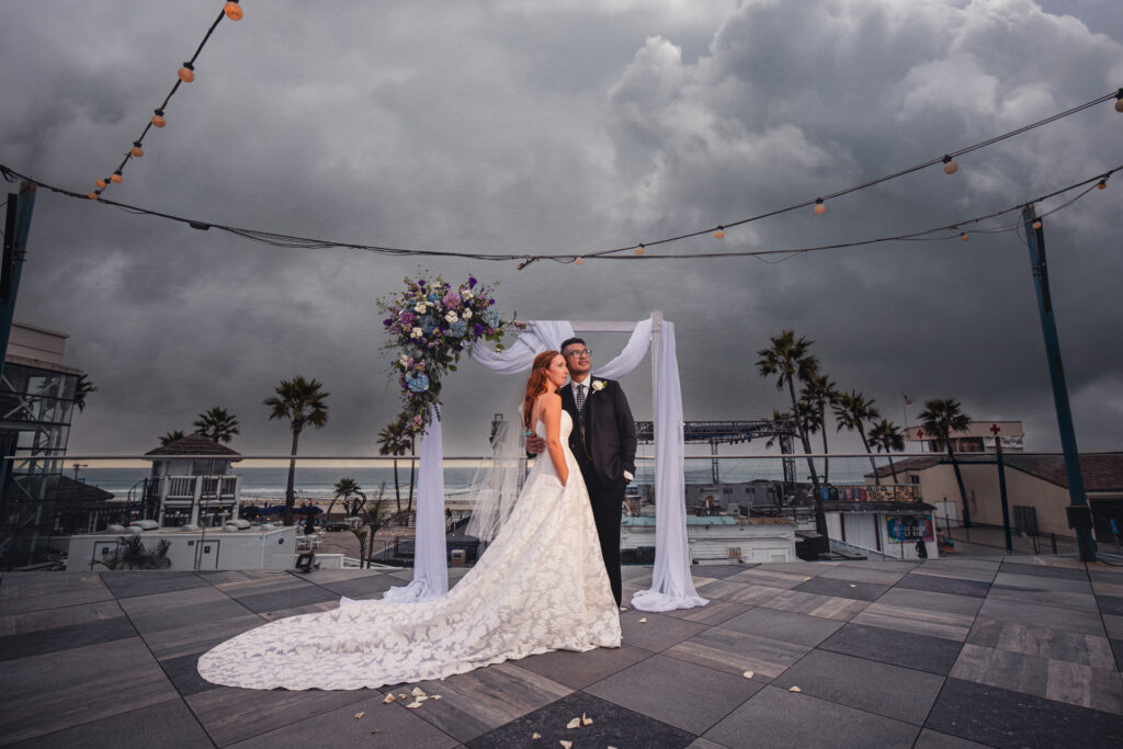 A bride in a vintage wedding dress and a groom embrace under their wedding floral arch at Belmont Park in Mission Beach.