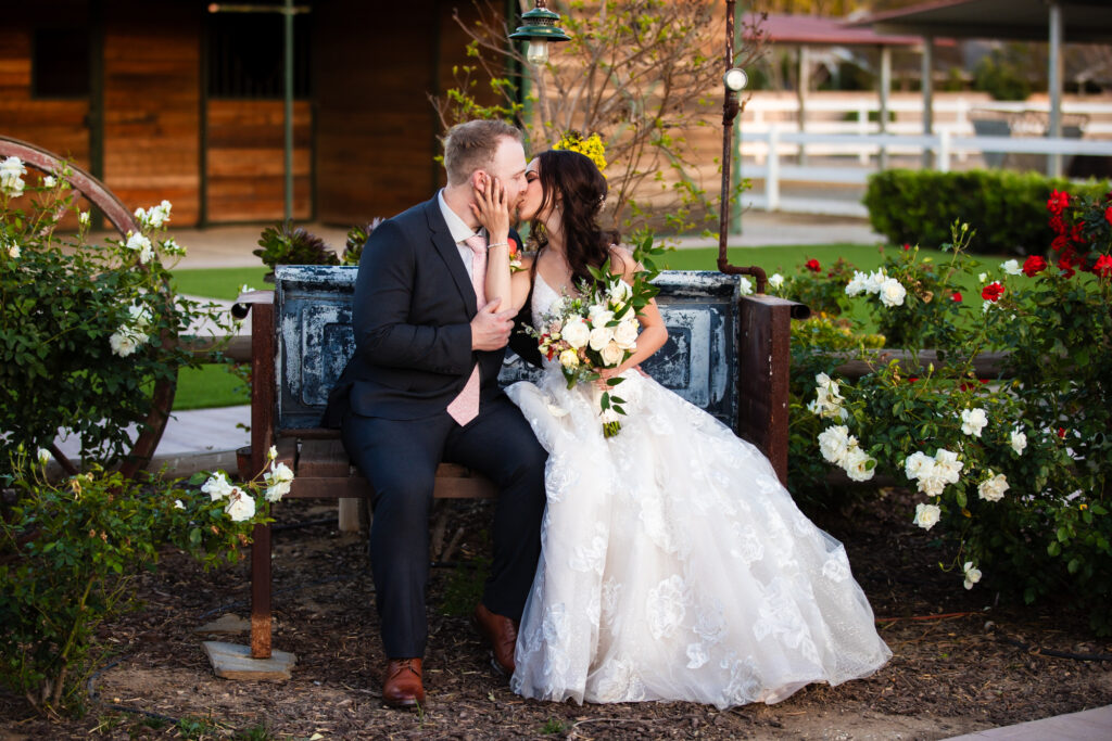 A groom and a bride holding her wedding bouquet share a kiss on a rustic bench on their rustic wedding day at The Farm in Norco, a Southern California wedding venue.
