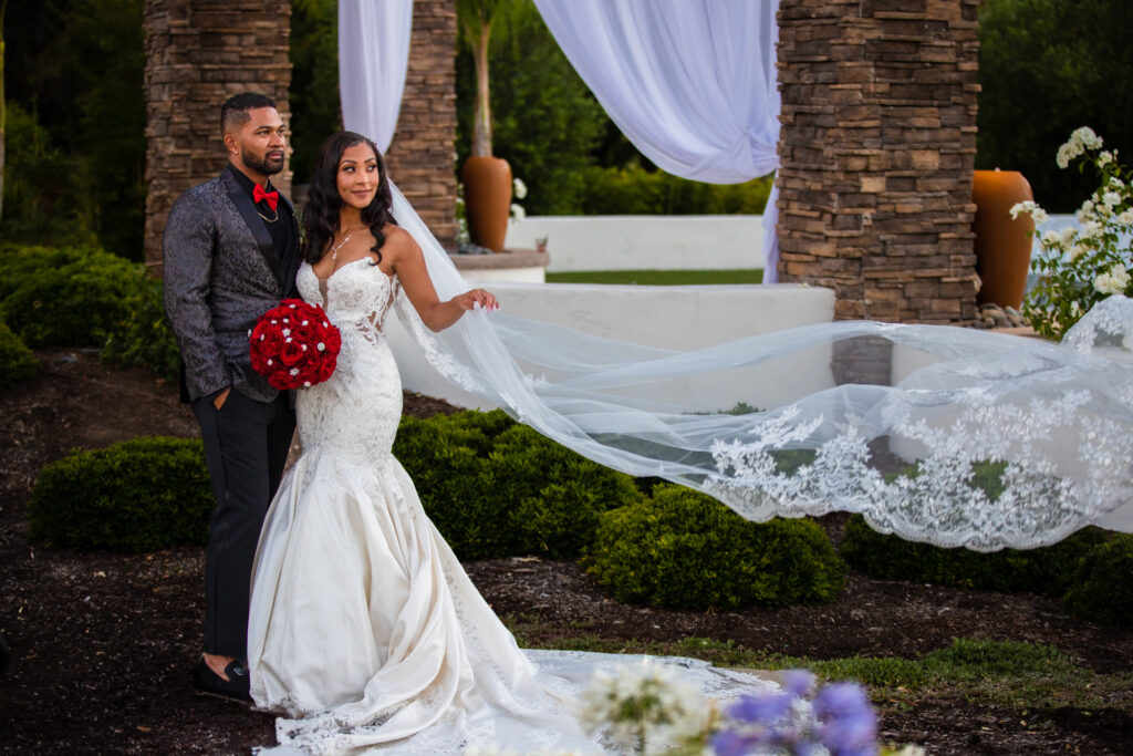 A groom with a custom tux with patterned tux jacket and a bride with a vibrant red bouquet and billowing veil embrace on their wedding day at Fallbrook Estate by Wedgewood Weddings in Southern California.