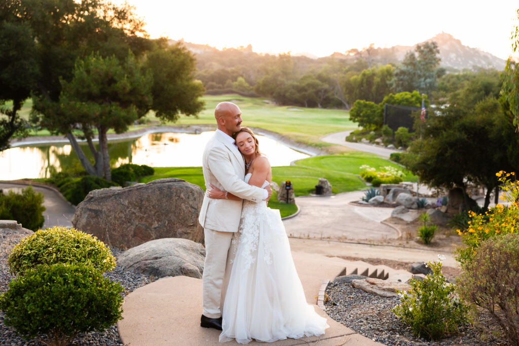 A fit, in-shape bride and groom share a quiet embrace overlooking greenery and a lake at Mt. Woodson Castle in Ramona, California.