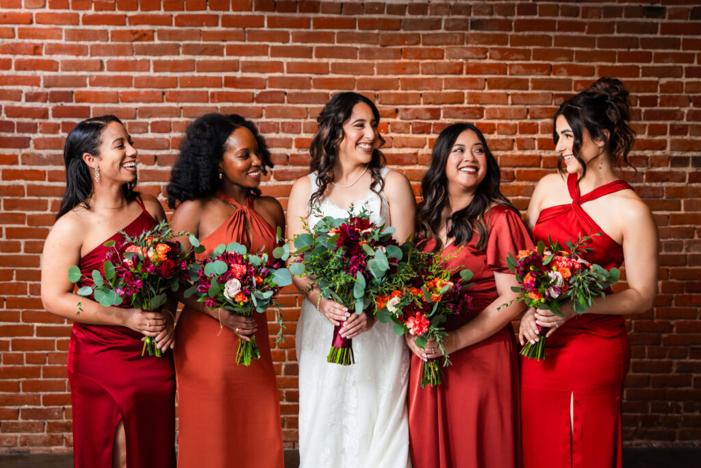 I bride and her bridesmaids in their red and burnt orange bridesmaid dresses smile and laugh in front of a brick wall at The Sinclair on G Street, a wedding venue in Downtown San Diego.