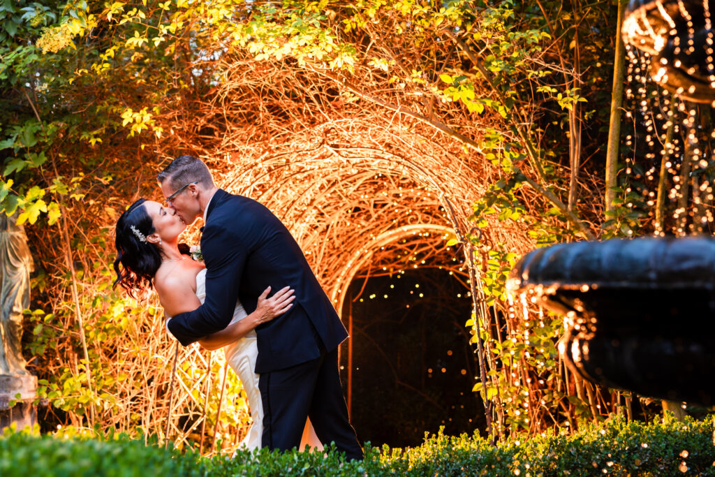 A happy bride and groom share a dip kiss by a fountain surrounded by greenery on their wedding day at Twin Oaks Garden House Estate in San Marcos, California.